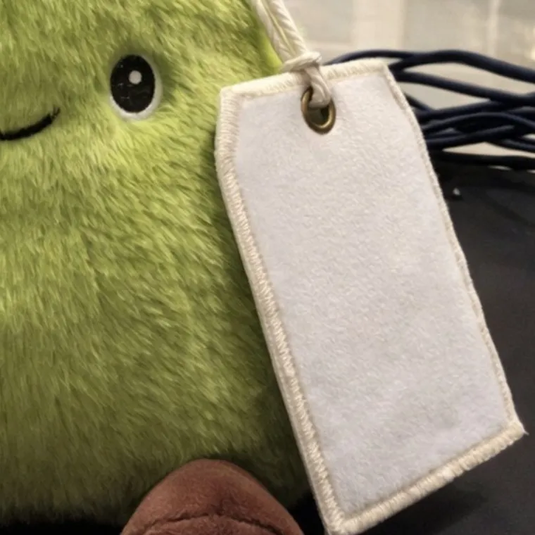 Close-up of the blank white fleece tag on Percy the Pear, showing the brass grommet, cord and blanket-stitched edge
